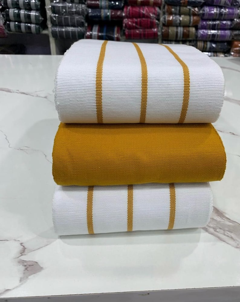 May include: Three rolls of fabric, one white with gold stripes, one solid gold, and one white with gold stripes. The fabric is folded and stacked on a white surface.