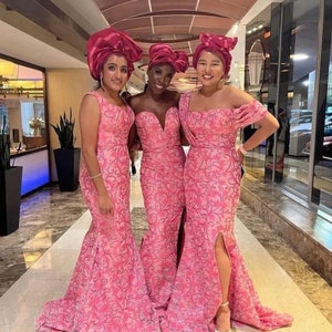 Puede incluir: Tres mujeres con vestidos de sirena de encaje rosa y turbantes a juego. Los vestidos tienen un estampado floral y una abertura lateral. Las mujeres est&aacute;n de pie en un pasillo con suelo de baldosas.