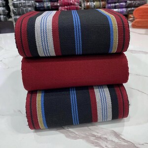 May include: A stack of three fabric rolls with striped patterns. The top roll is black with white and blue stripes, the middle roll is red, and the bottom roll is black with white, blue, and gold stripes.