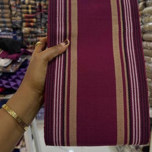 Può includere: Un tessuto piegato con una base bordeaux scuro, accentuato da strisce verticali beige, bianche e nere. Il tessuto ha una trama tessuta e le strisce creano un motivo simmetrico. Il tessuto è tenuto in mano, evidenziando il colore e il design.