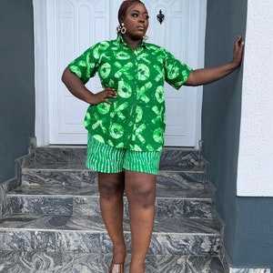 May include: A green two-piece outfit featuring a short-sleeved button-down shirt and matching shorts. The shirt and shorts have a repeating pattern of white shapes on a green background. The shorts have a striped pattern at the bottom. The outfit is worn with clear sandals.