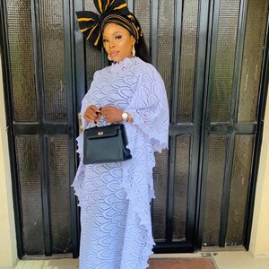 May include: A woman wearing a white lace dress with a black and yellow head wrap and a black handbag.