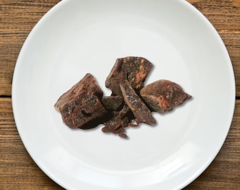 May include: A white ceramic plate holds several pieces of dark brown, irregularly shaped chunks. The pieces have a rough texture and varying shades of brown and reddish-brown. The plate is set against a wooden background.