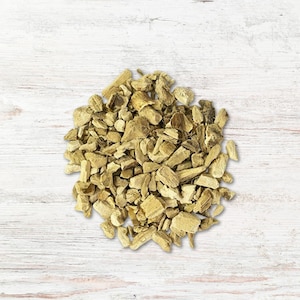 May include: A pile of chopped, dried, light brown ginger root pieces. The ginger pieces are irregularly shaped and vary in size. The background is a white wooden surface with visible wood grain.