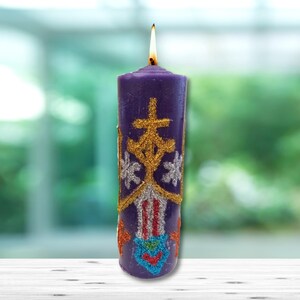 May include: A purple candle with a gold and silver glitter design. The candle has a flame on top and is sitting on a white wooden surface.