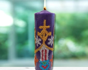 May include: A purple candle with a gold and silver glitter design. The candle has a flame on top and is sitting on a white wooden surface.