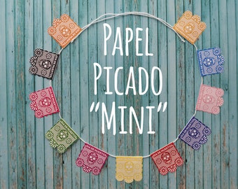 Può includere: Una ghirlanda di bandierine di carta piccata colorate con un designo a forma di teschio e il testo "Dia de Muertos" è appesa su uno sfondo di legno blu. Il testo "Papel Picado "Mini" è scritto in bianco al centro dell'immagine.