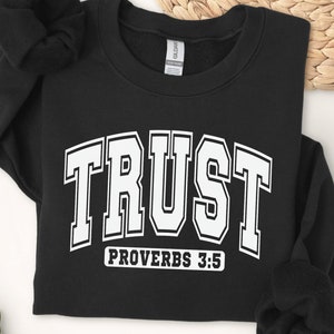 May include: Black sweatshirt with white text that reads "TRUST" in a collegiate font with "PROVERBS 3:5" underneath.