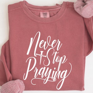 Puede incluir: Una sudadera color marrón rojizo con el texto "Never Stop Praying" en letras blancas. El texto incluye un pequeño símbolo de cruz.