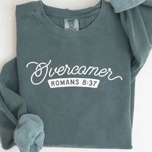 Könnte beinhalten: Ein blaugrüner Pullover mit dem Aufdruck "Overcomer Romans 8:37" in Weiß.