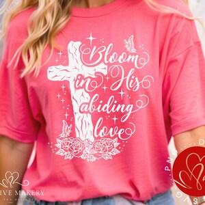 Può includere: Maglietta rosa con un design grafico bianco di una croce con il testo "Bloom in His abiding love" e farfalle.