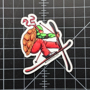 May include: A sticker featuring a green and red cartoon turtle wearing a red bandana and red pants, skiing down a slope. The turtle is holding ski poles and has a shell on its back. The sticker has the text "@COSMICMELLEN" on it.