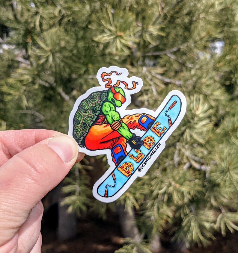 Puede incluir: Una pegatina verde, naranja y azul de una Tortuga Ninja Adolescente haciendo snowboard en una tabla de snowboard con forma de pizza. La tabla de snowboard tiene una cadena unida a ella y el texto "@cosmicmcillen" est&aacute; impreso en la parte inferior.