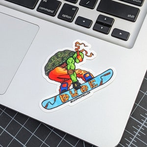 Puede incluir: Una pegatina con un personaje de las Tortugas Ninja de color verde, naranja y rojo haciendo snowboard en una tabla de snowboard con tem&aacute;tica de pizza. La tabla de snowboard tiene la palabra "DUDE" escrita en una fuente colorida. La pegatina tiene el nombre de Instagram del artista, @cosmicmellen, en la parte inferior.