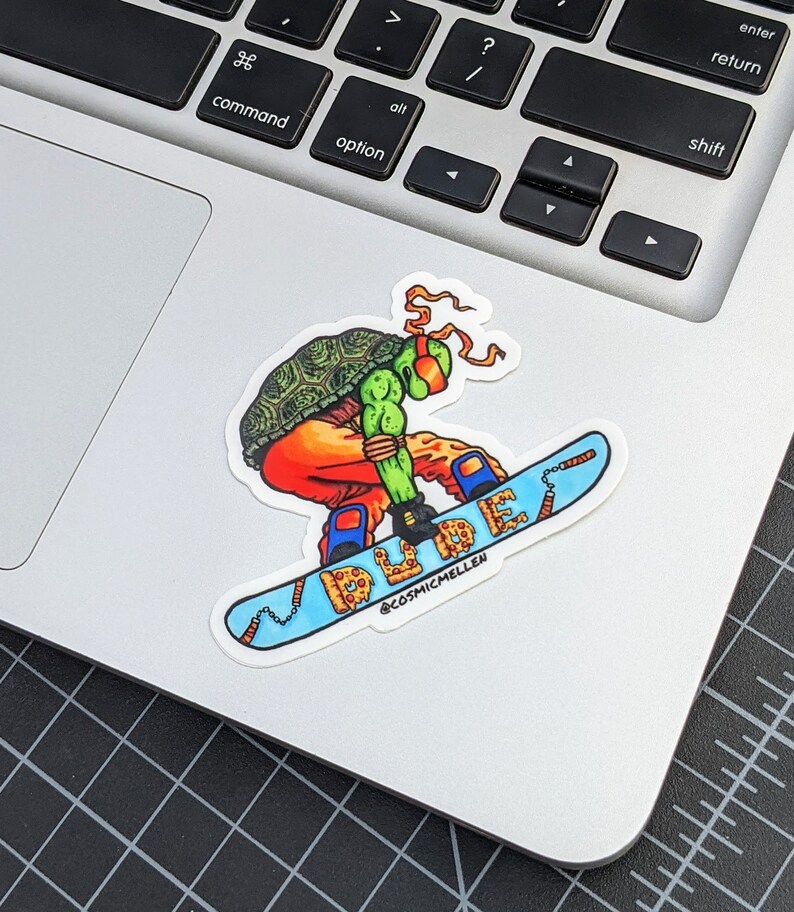 Puede incluir: Una pegatina con un personaje de las Tortugas Ninja de color verde, naranja y rojo haciendo snowboard en una tabla de snowboard con tem&aacute;tica de pizza. La tabla de snowboard tiene la palabra "DUDE" escrita en una fuente colorida. La pegatina tiene el nombre de Instagram del artista, @cosmicmellen, en la parte inferior.