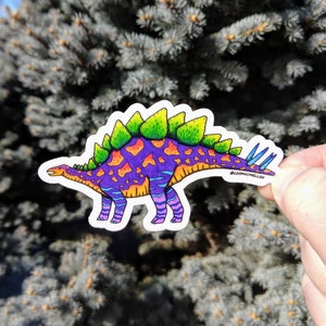 May include: A colorful sticker of a dinosaur with green spikes, purple and orange spots, and blue stripes. The dinosaur is standing on a white background. The sticker has the text "@cosmicmellen" on the bottom right corner.