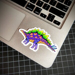 May include: A colorful sticker of a dinosaur with purple, green, orange, and blue accents. The dinosaur has spikes on its back and a long tail. The sticker is on a silver laptop.