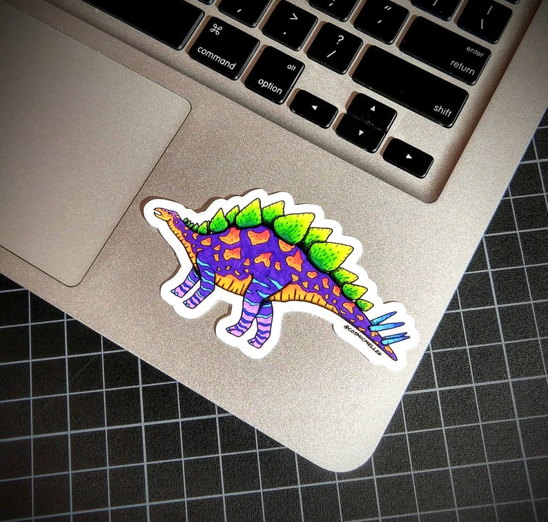 May include: A colorful sticker of a dinosaur with purple, green, orange, and blue accents. The dinosaur has spikes on its back and a long tail. The sticker is on a silver laptop.