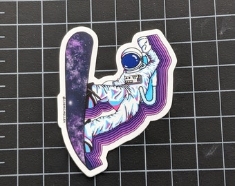 May include: A sticker of an astronaut snowboarding on a galaxy-themed snowboard. The astronaut is wearing a white spacesuit with a blue helmet and is surrounded by purple and blue lines.