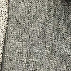 May include: Close-up of a textured gray fabric with a woven appearance. The fabric has a speckled pattern with small white and dark gray flecks. A knitted border in white and gray is visible on the left side, adding a contrasting texture.