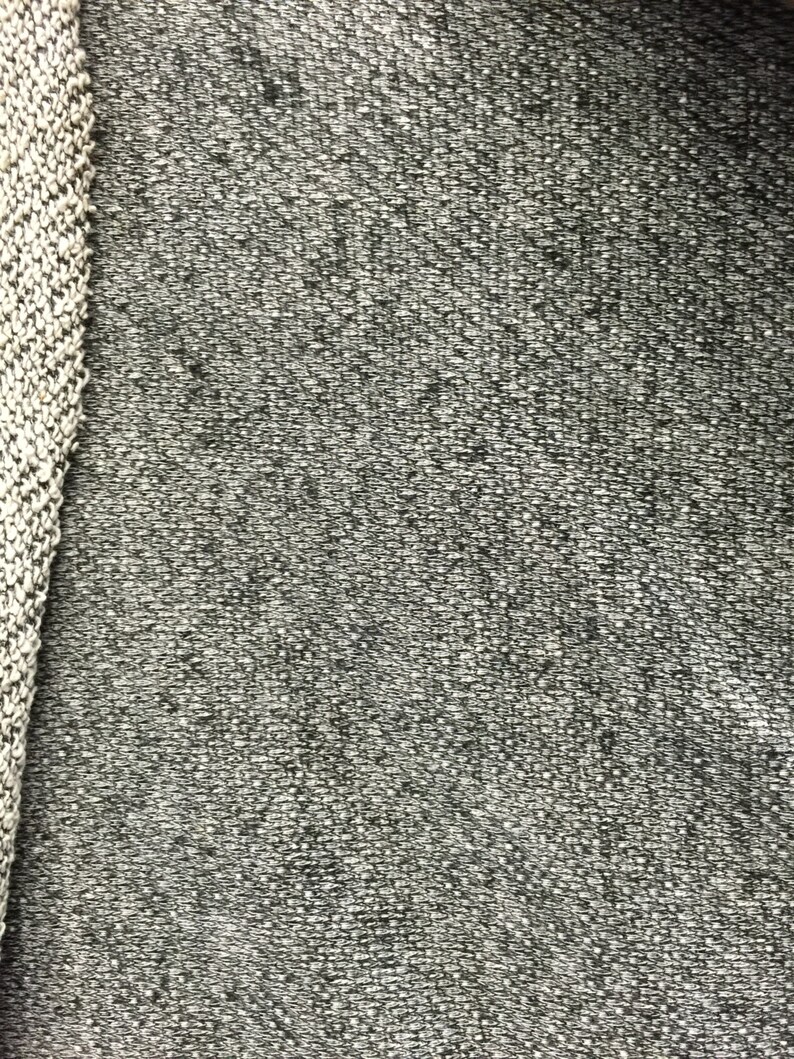 May include: Close-up of a textured gray fabric with a woven appearance. The fabric has a speckled pattern with small white and dark gray flecks. A knitted border in white and gray is visible on the left side, adding a contrasting texture.