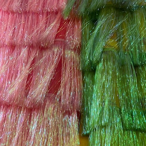 Könnte beinhalten: Nahaufnahme von Bündeln aus rosa und grünem, schillerndem Kunsthaar. Das Haar ist mit kleinen Bändern zusammengebunden. Das rosa Haar hat ein schimmerndes, glitzerndes Aussehen, während das grüne Haar einen hellen, lebendigen Farbton hat.