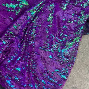 May include: Close-up of a purple fabric covered in iridescent sequins. The sequins have a mix of green and blue hues, creating a shimmering effect. The fabric is draped, showing folds and texture.