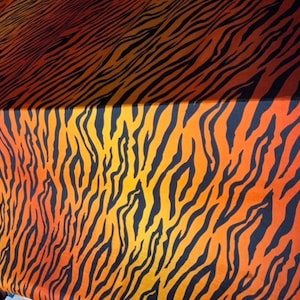 May include: Fabric with a tiger stripe pattern. The fabric is divided into two sections. The top section has a dark brown and black pattern. The bottom section has an orange and yellow gradient with black stripes.