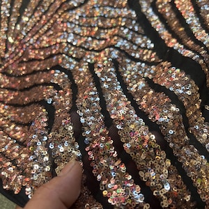 May include: Close-up of a fabric with a black mesh base, covered in shimmering copper-coloured sequins. The sequins are arranged in a striped pattern, creating a textured and visually dynamic surface. The fabric has a luxurious and glamorous appearance.