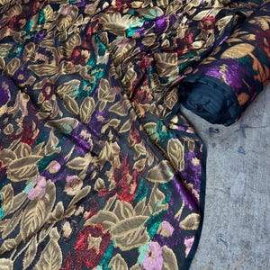 May include: Black fabric featuring a floral design in gold, red, purple, and green. The fabric is draped and rolled, highlighting the detailed pattern. The floral design includes leaves and flowers, creating a luxurious textile.