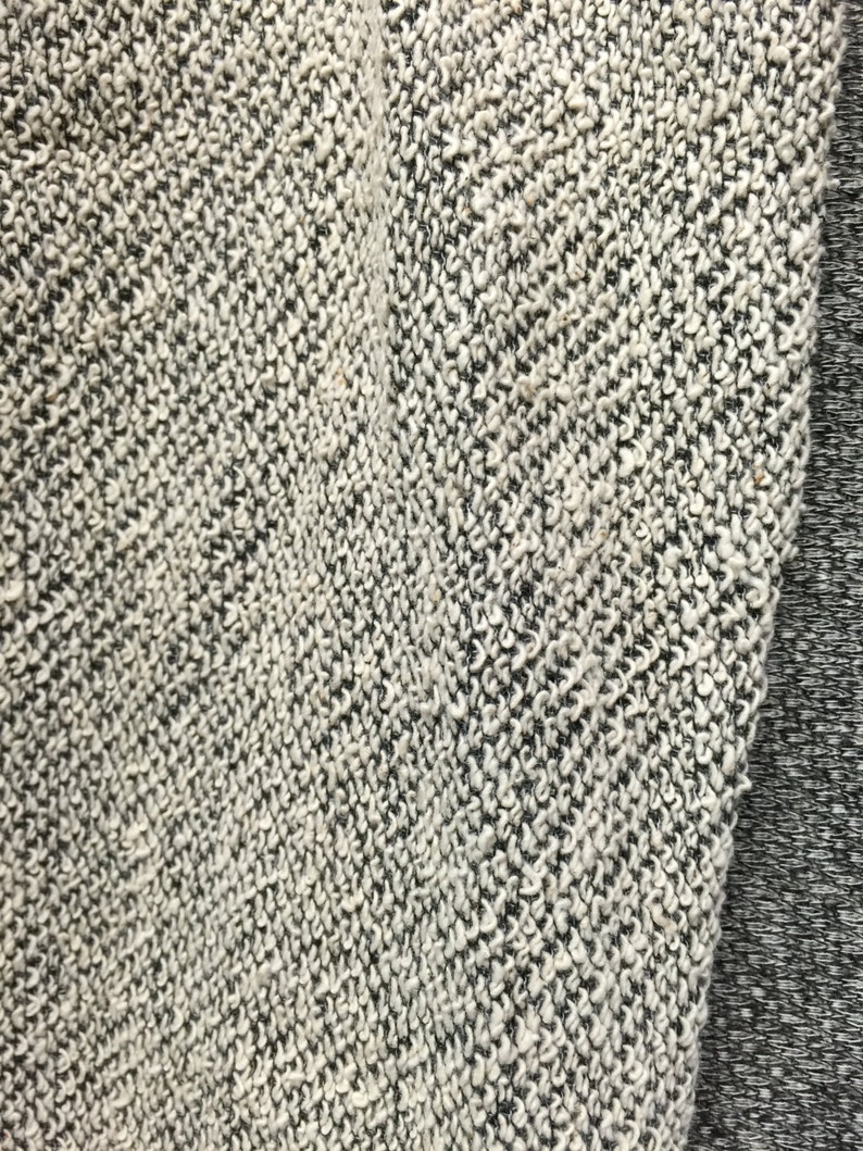 May include: Close-up of a textured fabric with a speckled pattern. The fabric is primarily off-white with black flecks, creating a heathered appearance. A section of solid gray fabric is visible on the right side, providing contrast.