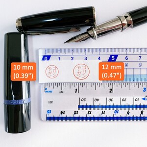 May include: A close-up of a ruler with two black fountain pens. The ruler is marked in inches and centimeters. The pens are both black with silver accents. The ruler is marked with the text "モーガン" in Japanese. The pens are 10 mm (0.39 inches) and 12 mm (0.47 inches) in diameter.