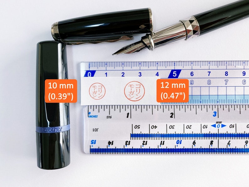 May include: A close-up of a ruler with two black fountain pens. The ruler is marked in inches and centimeters. The pens are both black with silver accents. The ruler is marked with the text "モーガン" in Japanese. The pens are 10 mm (0.39 inches) and 12 mm (0.47 inches) in diameter.