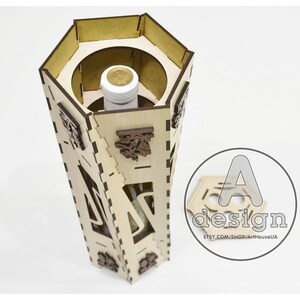 May include: A wooden hexagonal box with intricate laser cut designs and a bottle inside. The box is made of light brown wood and has a dark brown lid. The lid is open and the bottle is visible inside. The box is sitting on a white surface. The text "A design" is visible on the lid.