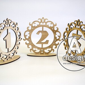 May include: Three wooden table number holders with ornate frames. The numbers 1, 2, and 3 are cut out of the wood and placed inside the frames.