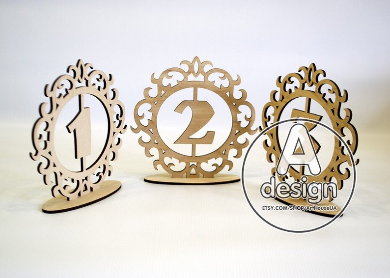 May include: Three wooden table number holders with ornate frames. The numbers 1, 2, and 3 are cut out of the wood and placed inside the frames.