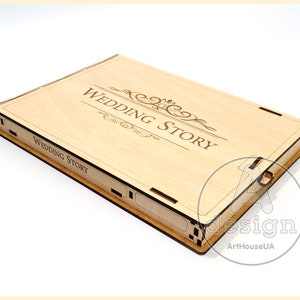 May include: A wooden box with a hinged lid, engraved with the words "Wedding Story" and a decorative scroll design. The box is made of light wood and has a natural finish.