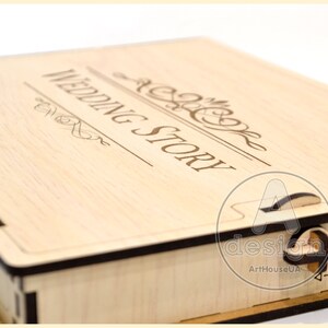 May include: A wooden box with a hinged lid. The lid is engraved with the words "Wedding Story" in a decorative font. The box has a black trim and a small latch on the side.