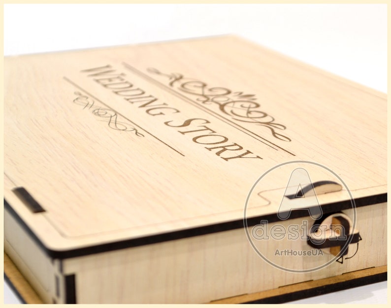 May include: A wooden box with a hinged lid. The lid is engraved with the words "Wedding Story" in a decorative font. The box has a black trim and a small latch on the side.