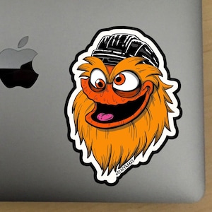 Peut inclure: Un autocollant représentant un monstre orange et brun de dessin animé portant un chapeau noir et blanc. Le monstre a un large sourire aux dents et regarde vers la droite. L'autocollant est sur un ordinateur portable argenté.