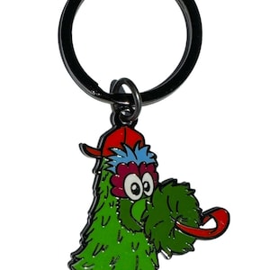 May include: A green, red, and blue keychain featuring a cartoon character with a red hat and a long, red tongue. The keychain has a black metal ring and a small loop connecting the charm to the ring. The character has large eyes and a green, feathery body.