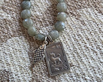 May include: A beaded necklace featuring faceted, gray-green beads. A silver pendant with a fish and a rectangular charm with a dog design hangs from the necklace. The necklace is displayed on a textured, neutral-colored surface.