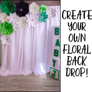 May include: A white backdrop with green and black paper flowers. The backdrop is decorated with a stack of blocks that spell out "BABY" in green letters.