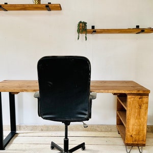 May include: A black office chair sits in front of a wooden desk with a shelf. The desk has a natural wood finish and black metal legs. There are two wooden shelves on the wall behind the desk.