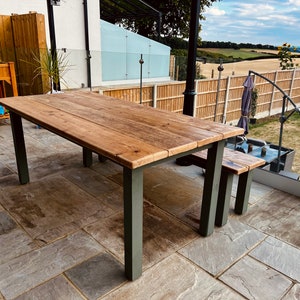 May include: A wooden outdoor dining table with a natural wood finish and dark grey metal legs. The table is set on a patio with stone tiles.