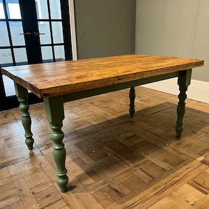 May include: A rectangular dining table featuring a natural wood grain tabletop with a warm brown finish. The table is supported by four turned legs painted in a matte sage green colour, showcasing a classic design with a smooth, rounded silhouette.