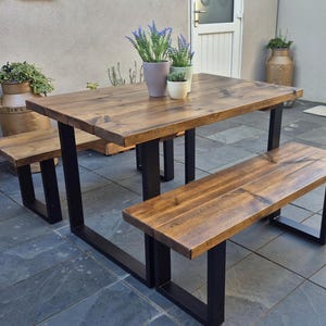 May include: A garden dining set featuring a rectangular wooden table and two benches. The table and benches have a dark wood finish and black metal legs. The set is surrounded by potted plants, creating a natural aesthetic.
