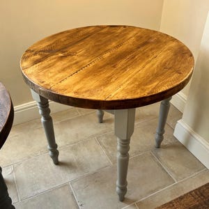 May include: A circular wooden table with a distressed finish. The tabletop is approximately 76 cm in diameter and features a natural wood grain pattern. The legs are painted a light grey colour, contrasting with the warm tones of the tabletop.