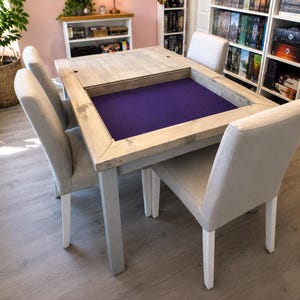 May include: A rectangular wooden gaming table with a light grey finish and a recessed purple felt playing surface. The table features a removable wooden top section and is surrounded by four upholstered chairs with light beige fabric and white legs.