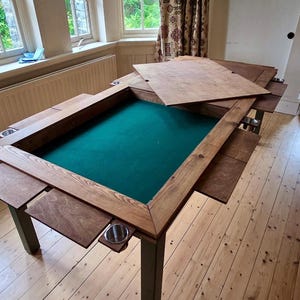 May include: A rectangular wooden gaming table featuring a recessed green felt surface, a thick natural wood frame, and removable wooden top panels. The table includes integrated metal cup holders attached to the sides and sturdy legs.